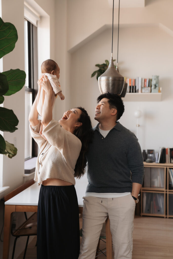 Natural light in-home family moment with mother lifting newborn baby near a window while father smiles, candid lifestyle newborn photography in a modern, cozy apartment.